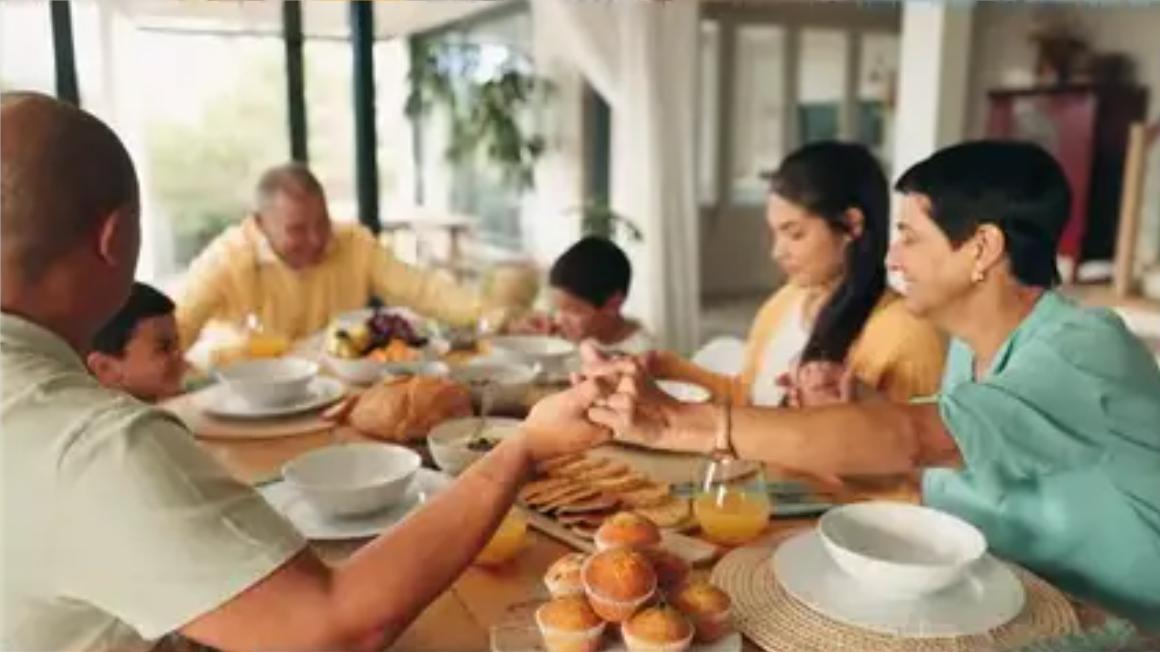 A multi-generational family holds hands together at the breakfast table
